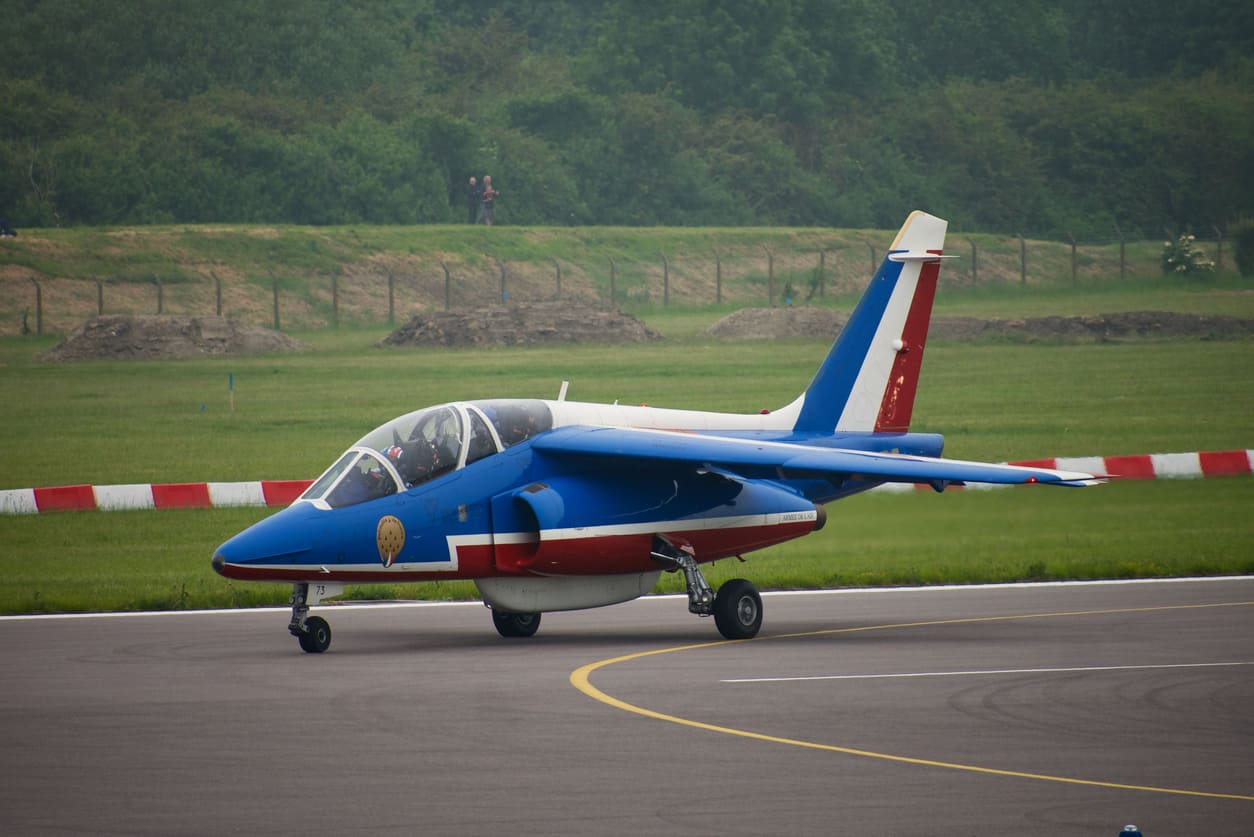 A vibrant aircraft is seen taxiing on the runway at Cambridge Airport, surrounded by green fields and a clear backdrop.