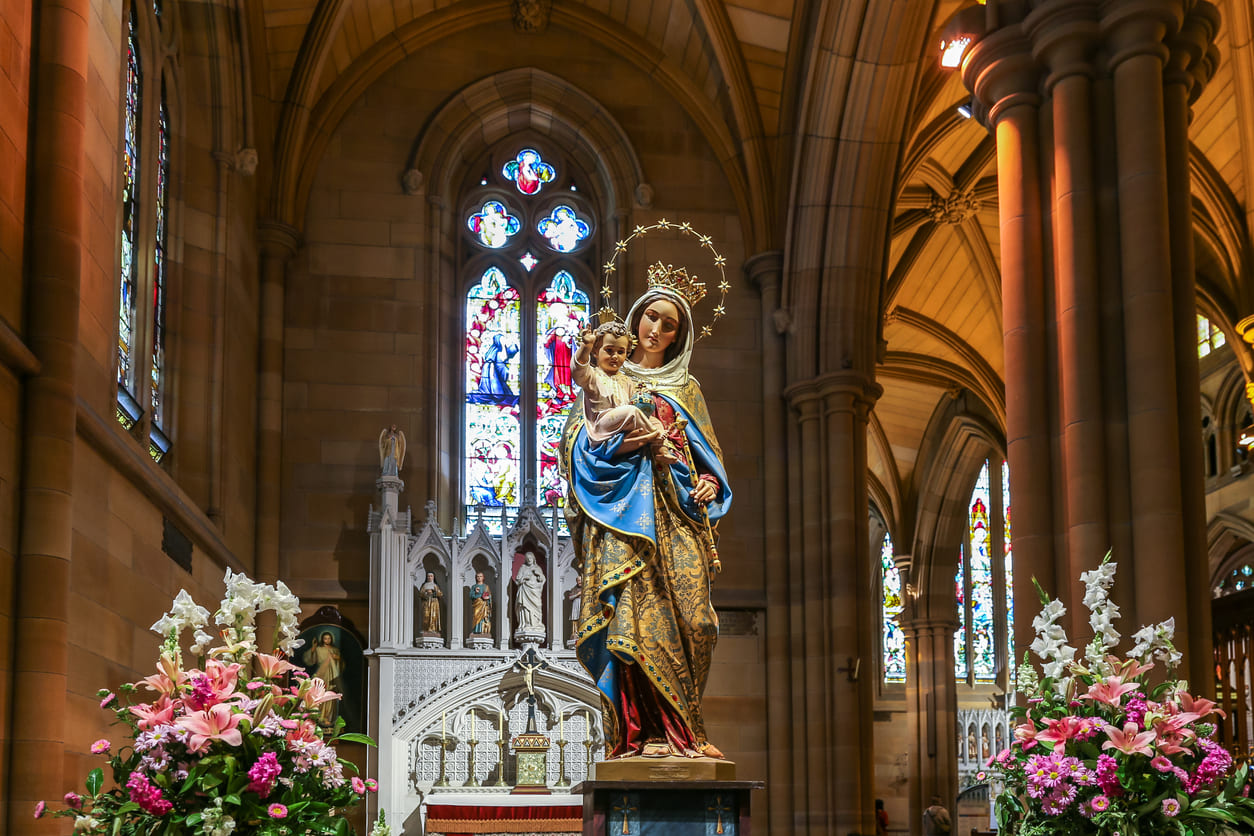 A richly decorated statue of the Virgin Mary with Child stands in a church, surrounded by flowers and stained glass, symbolizing devotion on Assumption Day.