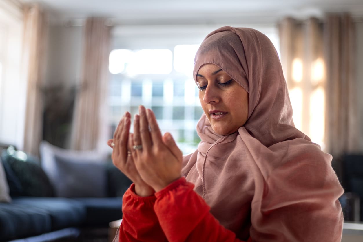 A Muslim woman in a pink hijab, seated indoors with hands raised in prayer, reflecting a moment of deep spiritual devotion.