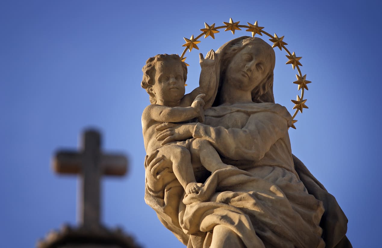 A serene statue of the Virgin Mary holding baby Jesus, crowned with stars, set against a clear blue sky and a blurred cross in the background.