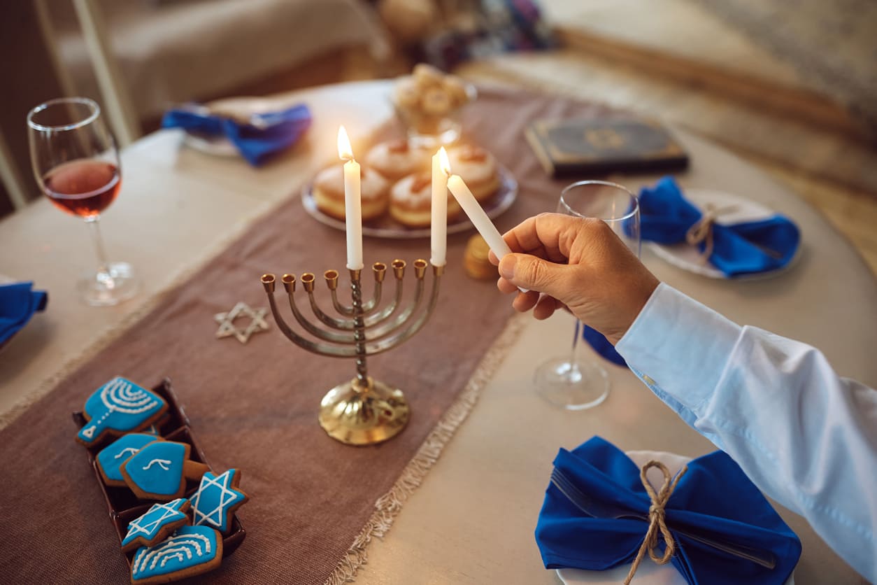 A festive table is set as a person lights the first candle on the menorah, surrounded by Hanukkah treats and symbols of the holiday.