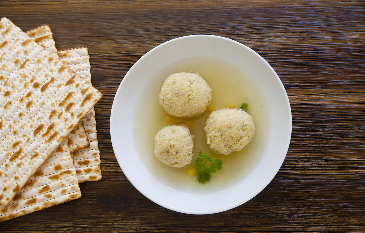 A bowl of matzo ball soup sits beside stacked matzah on a wooden table, reflecting the traditional flavors of Passover's first day.
