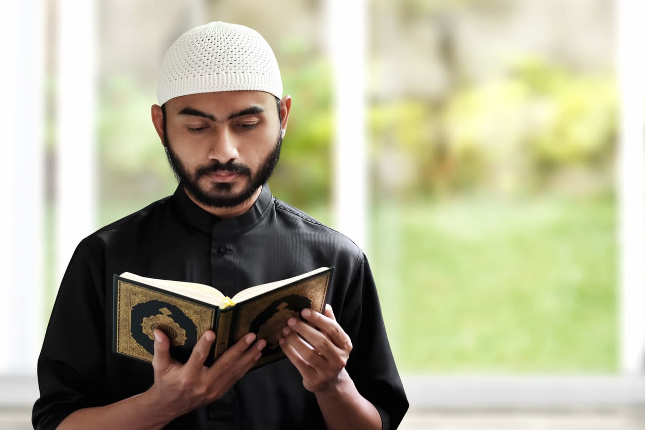 A man in traditional attire deeply focused on reading the Quran, standing in a calm, sunlit indoor setting.