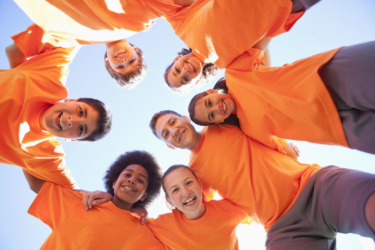 A joyful group of diverse children and an adult in orange shirts, standing in a circle and smiling down at the camera under a clear blue sky.