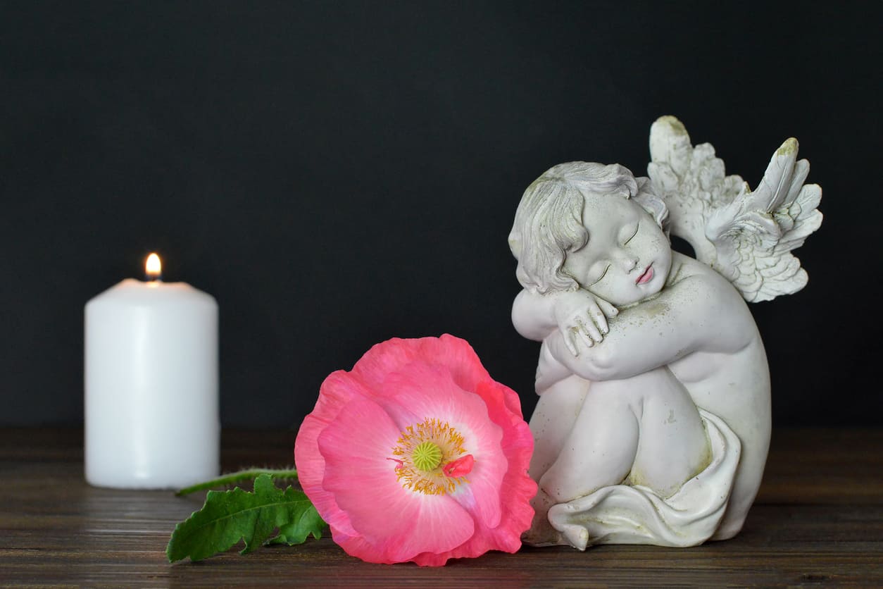 A peaceful scene with a lit candle, a sleeping angel statue, and a pink flower, symbolising remembrance and tenderness.