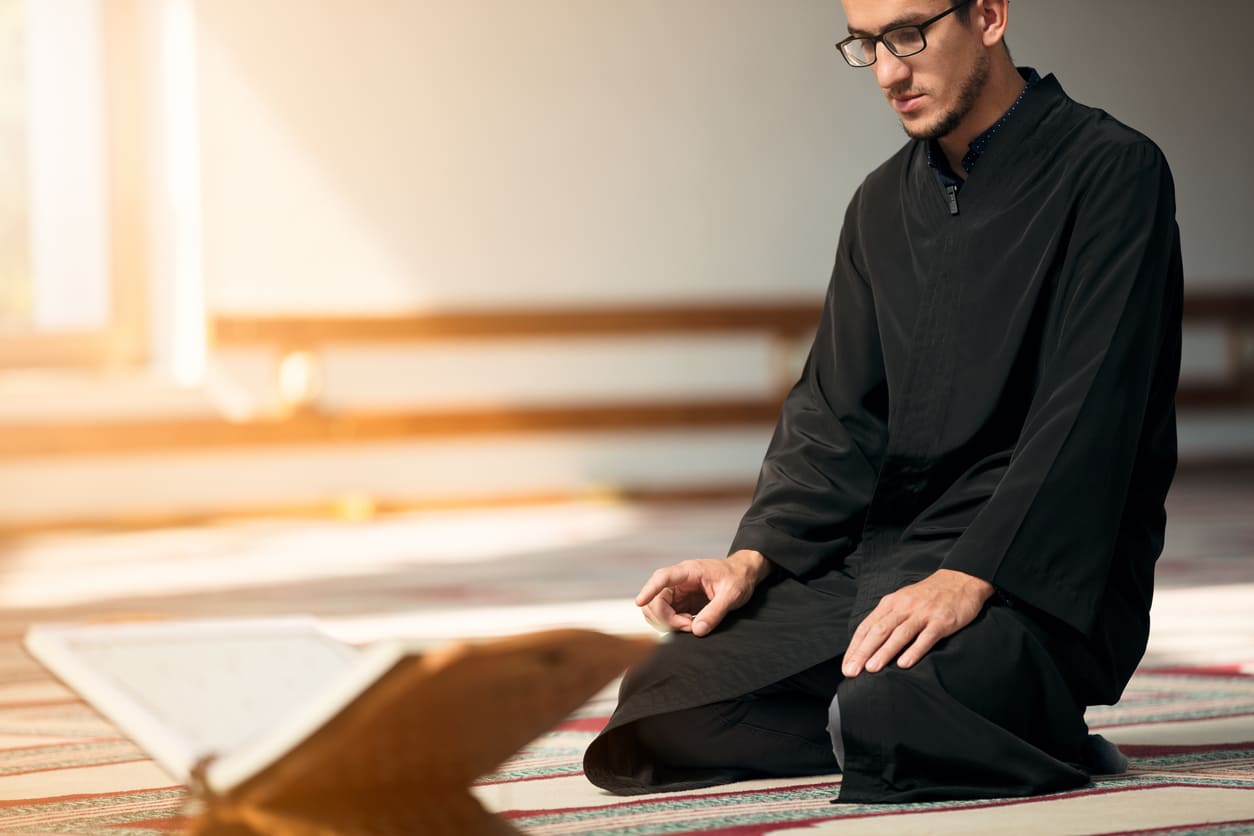 A man seated in prayer inside a mosque, deeply focused with the Quran open in front of him, reflecting a moment of devotion during Milad Un Nabi.