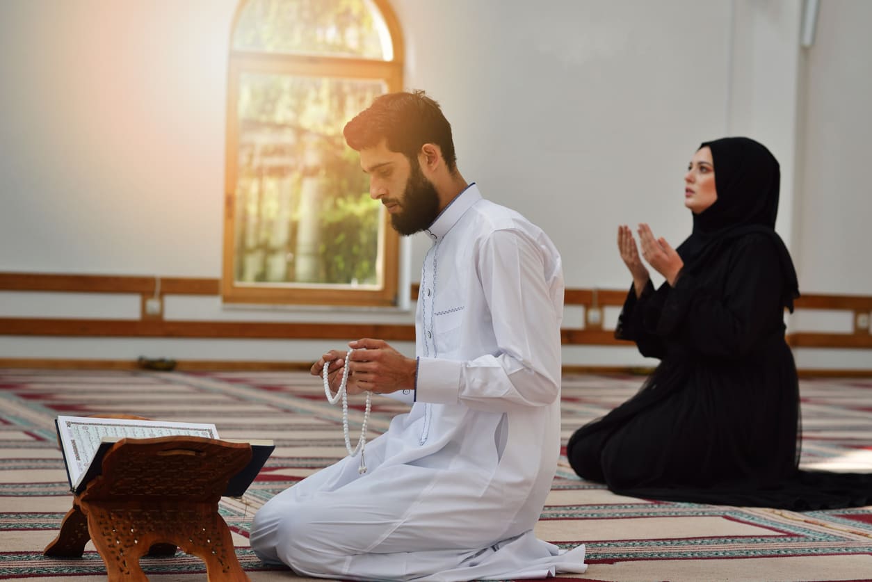 A man and woman praying inside a mosque, with the man holding prayer beads and the Quran placed before him, reflecting a moment of devotion during Muharram.