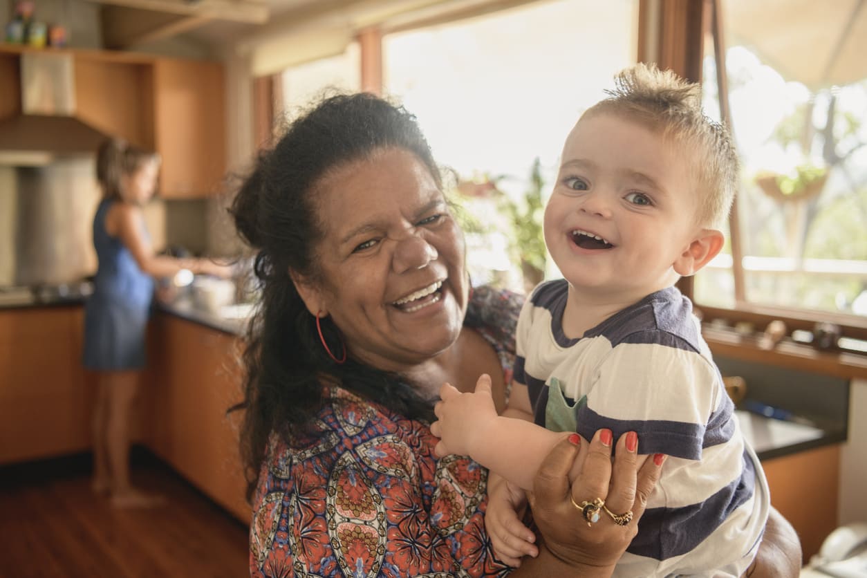 An Aboriginal woman joyfully holding a smiling child in a bright, homey setting, capturing warmth and connection across generations.