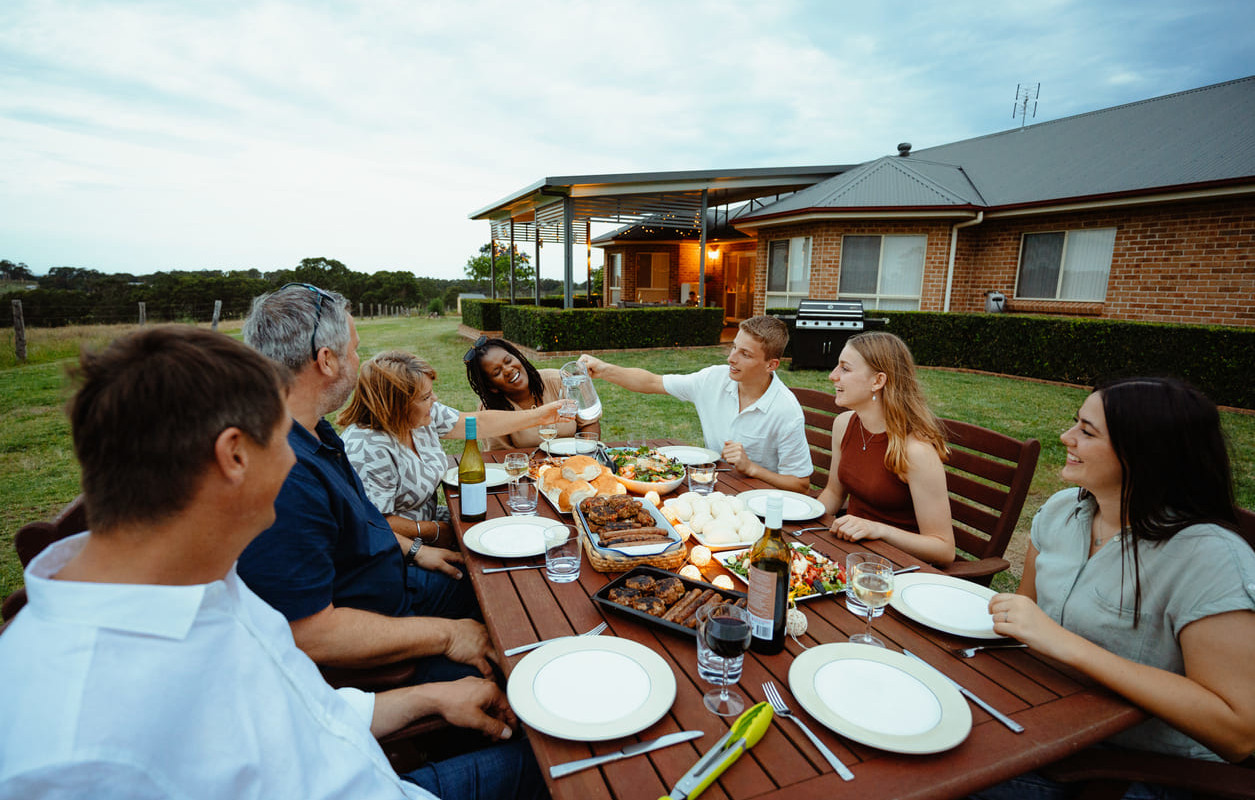 A cheerful group of people gathered around an outdoor table, sharing food and enjoying each other's company in a backyard setting.