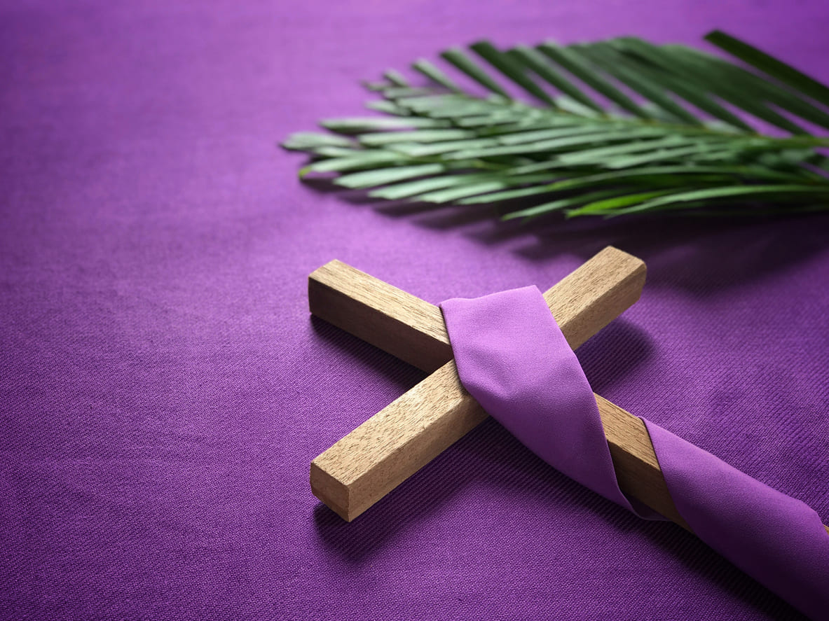 A wooden cross draped in purple cloth beside palm leaves, symbolizing the spirit and meaning of Palm Sunday.