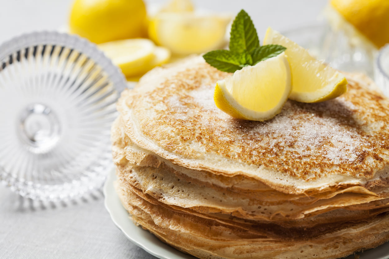 A close-up of thin, golden pancakes topped with lemon wedges and sugar, set against a bright table, reflecting the classic Pancake Day tradition.