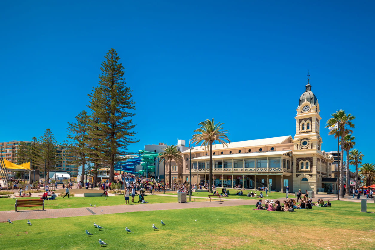 A sunny day at Glenelg with people gathered on the lawns near historic buildings, capturing the relaxed atmosphere of Proclamation Day.