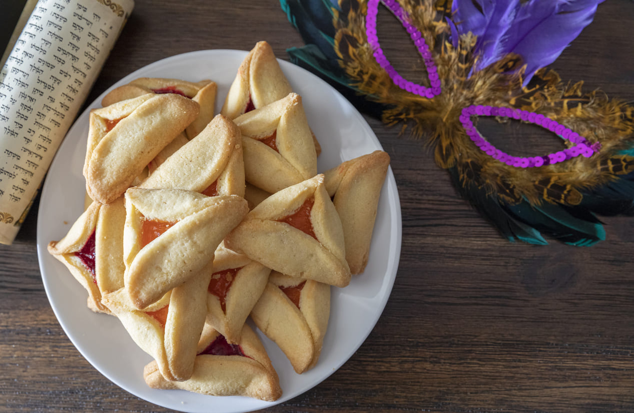 A plate of colorful hamantaschen sits beside a decorative mask and Megillah scroll, symbolizing the festive and meaningful traditions of Purim.