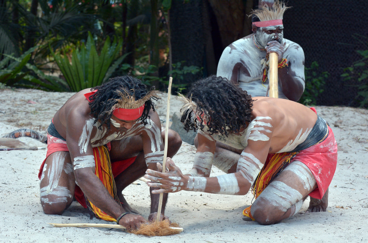 Aboriginal men in traditional attire performing fire-making and playing the didgeridoo as part of a cultural display in Queensland, Australia.