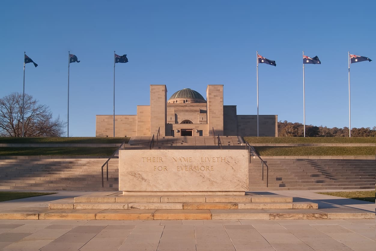 The Australian War Memorial in Canberra, framed by national flags and steps leading to a solemn stone inscription.