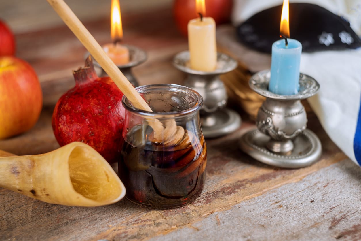 A close-up of Rosh Hashana symbols, including a shofar, honey jar, pomegranate, and lit candles, arranged on a rustic wooden table.