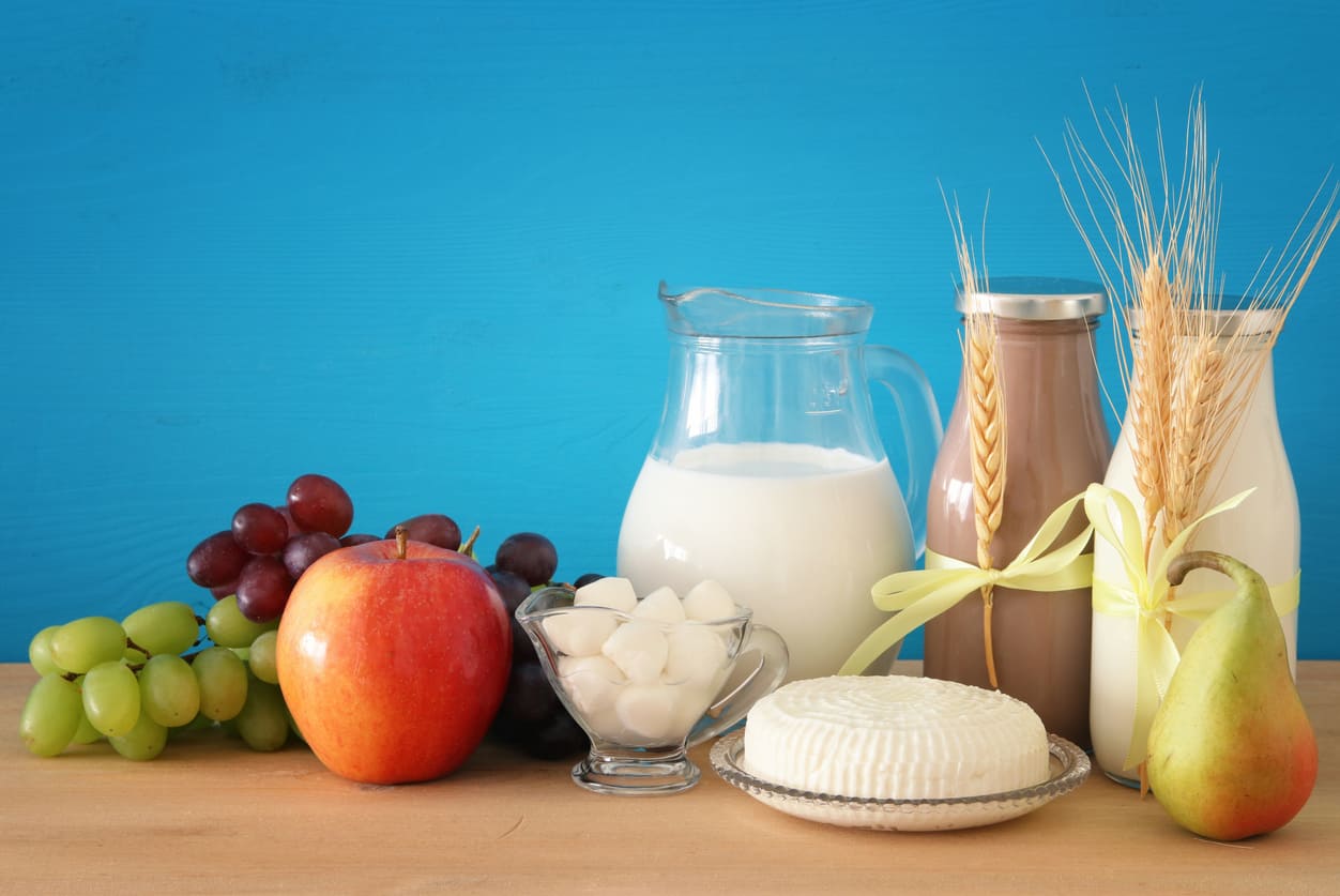 A festive arrangement of milk, cheese, fruits, and wheat sits against a bright blue background, reflecting Shavuot's harvest and dairy traditions.