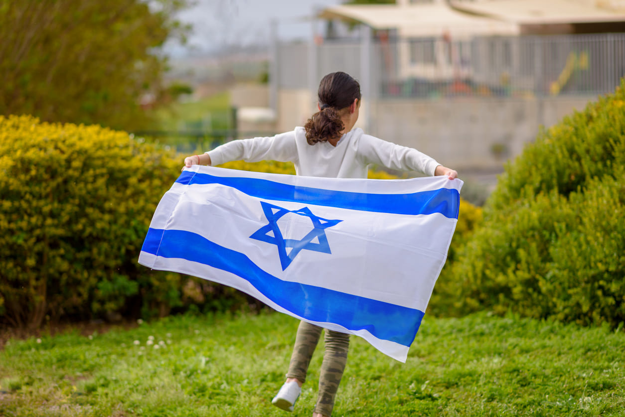 A young girl runs through a green garden holding the Israeli flag, symbolizing freedom and celebration on Yom Ha'atzmaut.