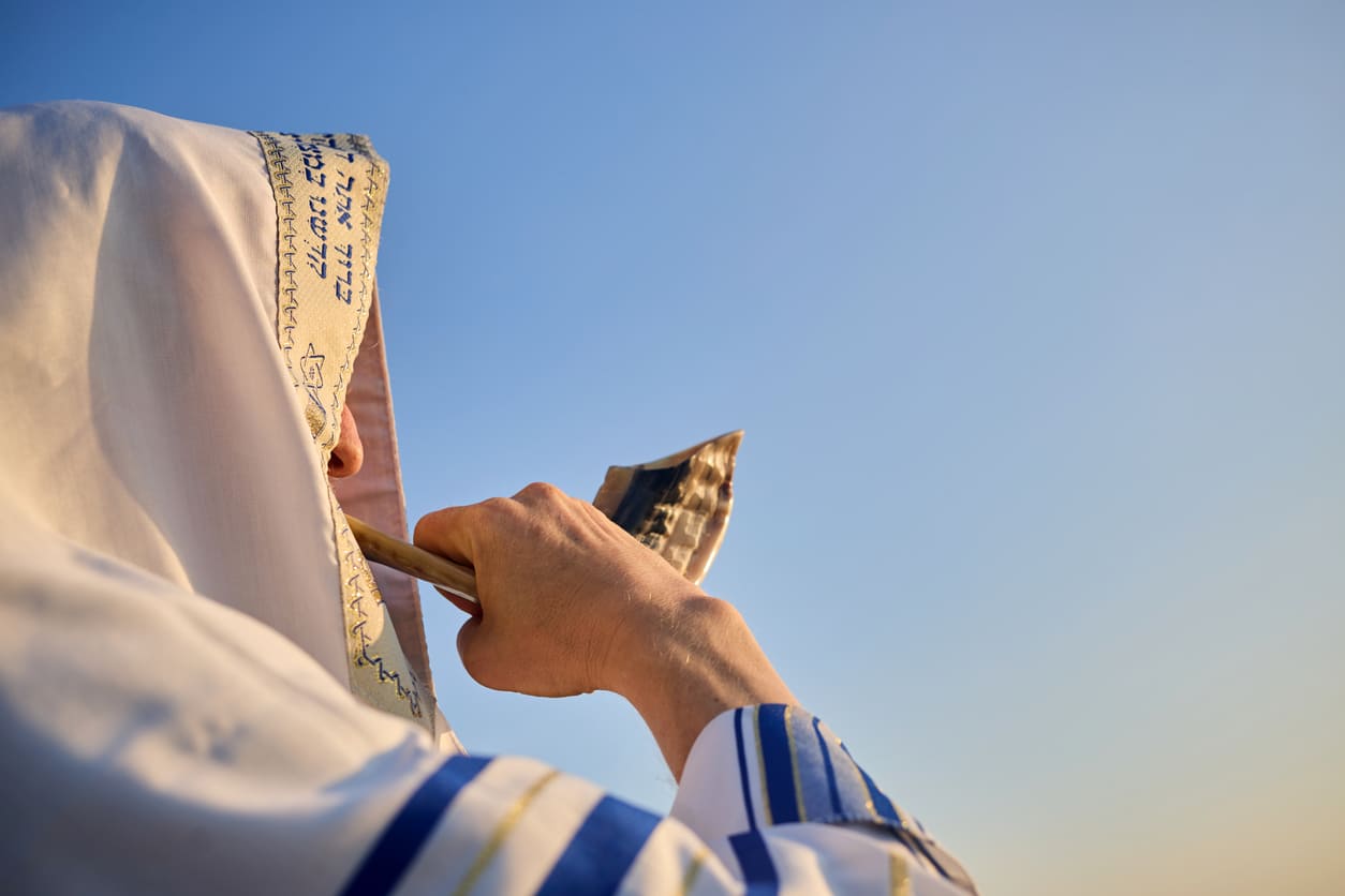 A person draped in a prayer shawl blows the shofar against a clear blue sky, capturing a sacred moment of Yom Kippur.