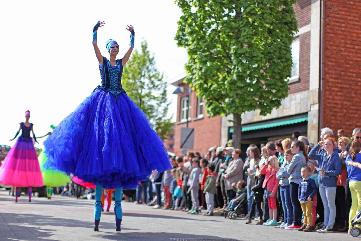 A lively scene from Belgium's annual May Day parade, featuring stilt performers in vibrant costumes entertaining a gathered crowd.