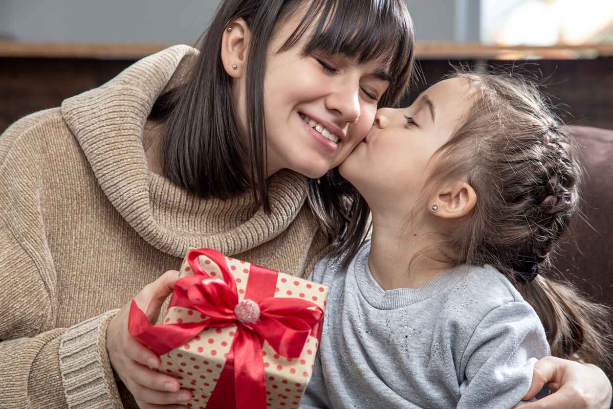 A touching Mother's Day scene of a young girl kissing her smiling mother while presenting a beautifully wrapped gift.