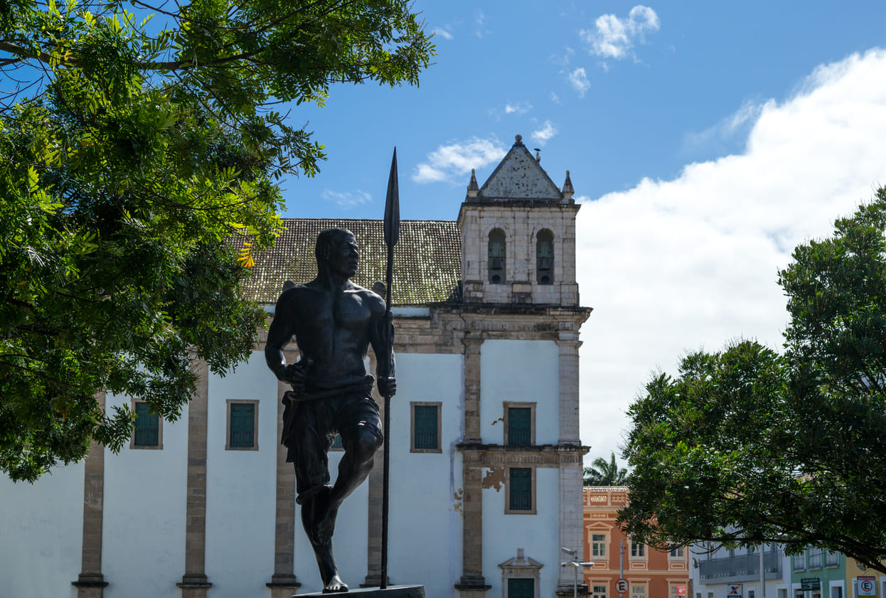  a striking statue of Zumbi dos Palmares holding a spear, set against a historic colonial building and framed by trees under a bright blue sky.