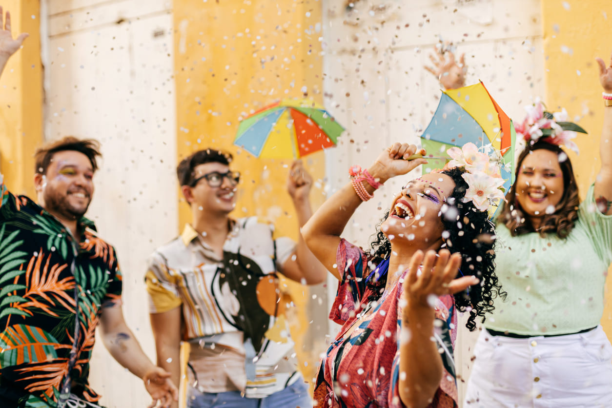 A lively group of people dressed in colorful outfits dance and smile under falling confetti, holding small Carnival umbrellas.