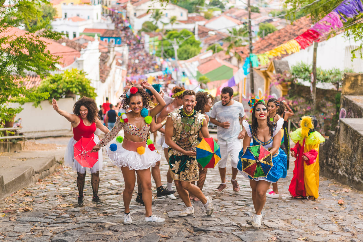 A joyful group of dancers in colorful costumes and frevo umbrellas parades up a cobblestone street, surrounded by festive decorations and cheering crowds.