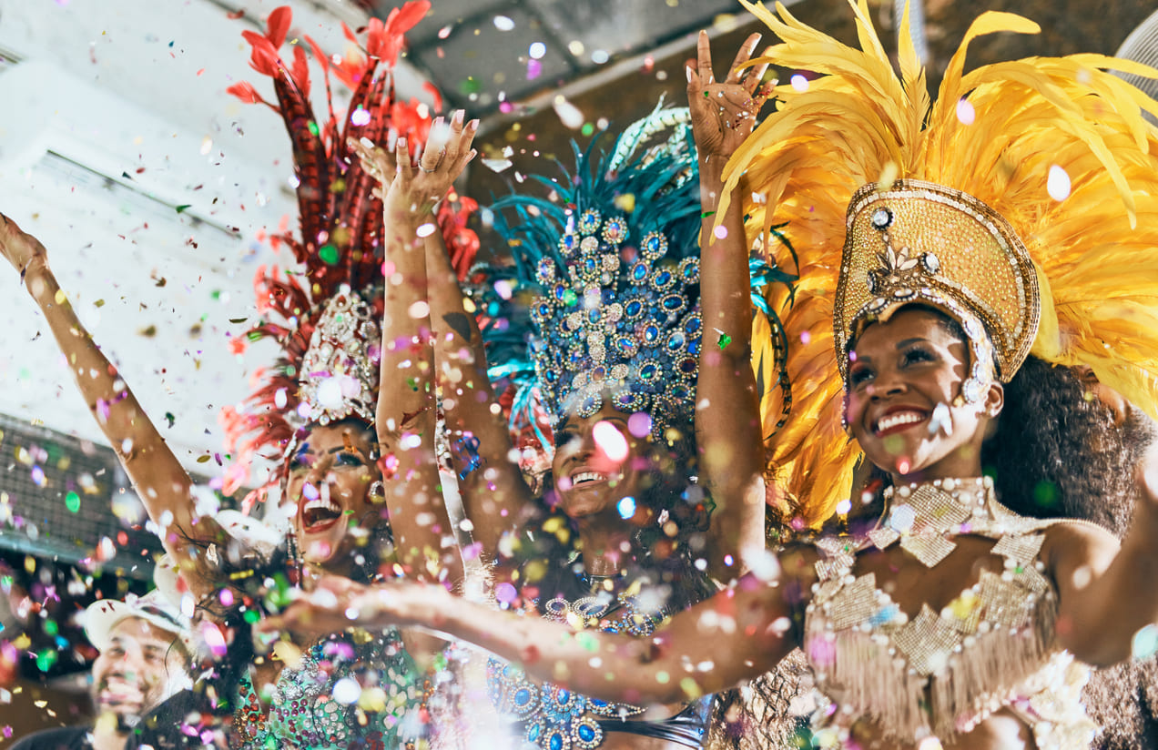 Three samba dancers in elaborate feathered costumes smile and raise their hands as confetti rains down, creating a festive and colorful scene.