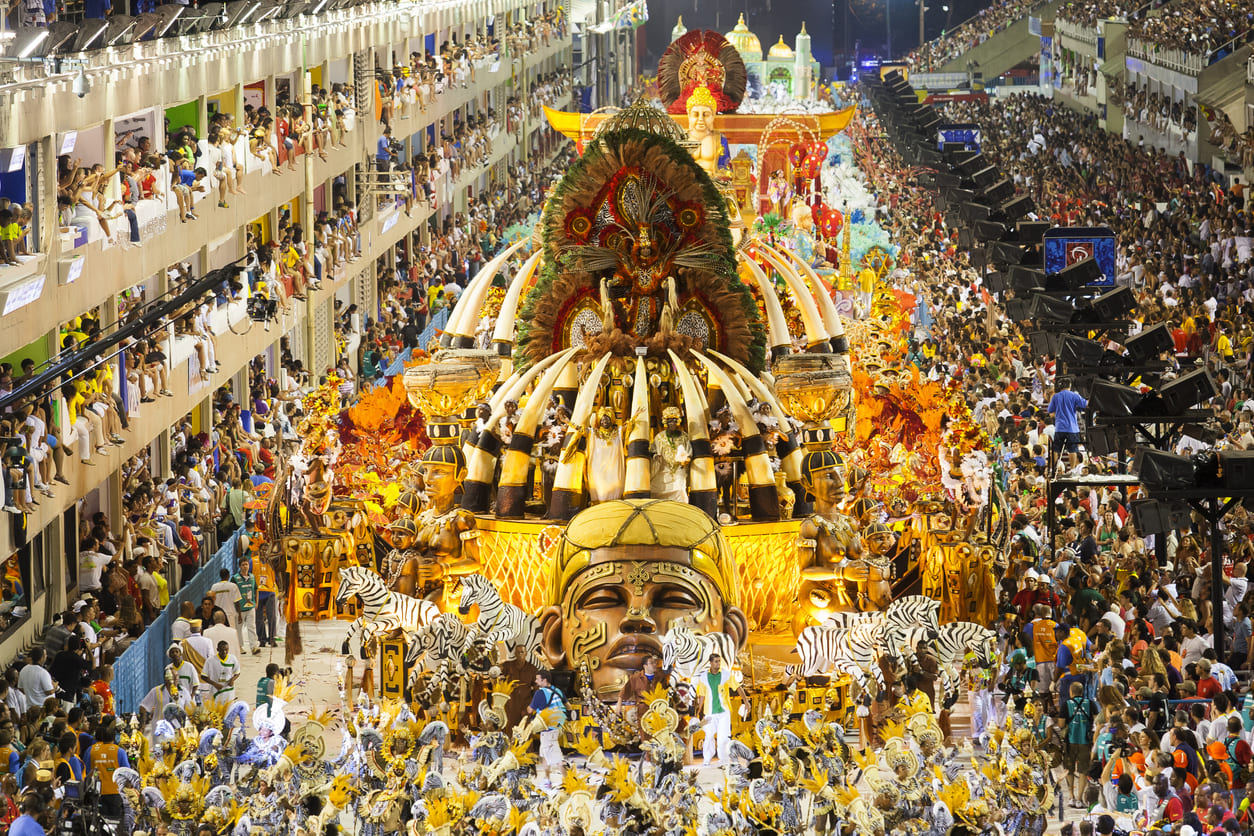 A massive, colorful float moves through a packed Sambadrome as samba dancers and performers in elaborate costumes entertain thousands of spectators.