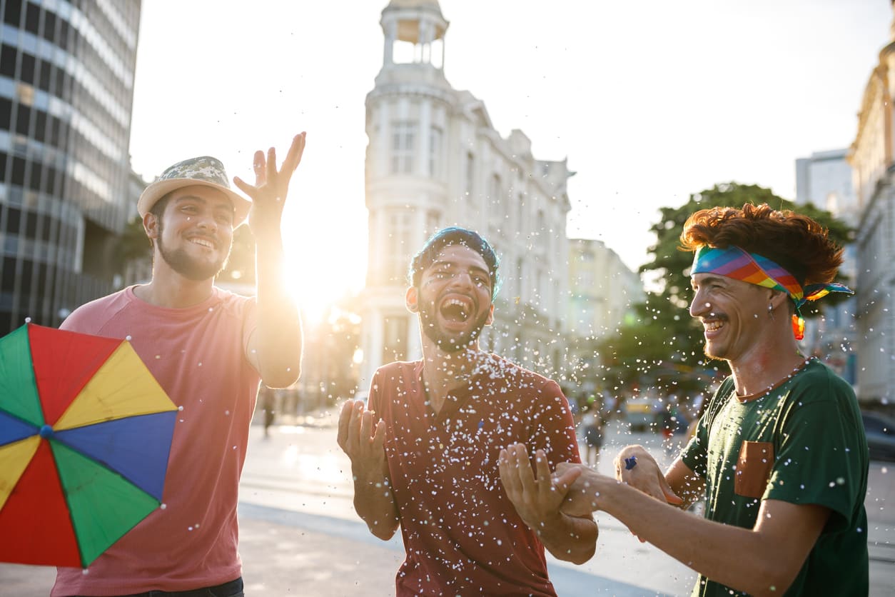 Three friends laugh and play with confetti on a sunny street, dressed in festive colors and surrounded by a cheerful Carnival atmosphere.