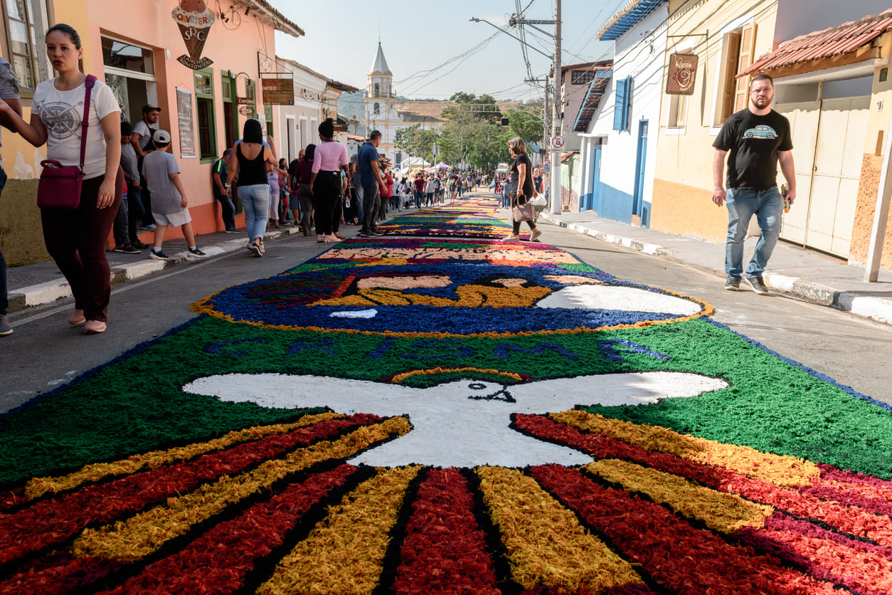 A vibrant street in Santana de Parnaíba, São Paulo, adorned with a colorful sawdust carpet for the Corpus Christi procession, with people walking alongside the artwork.