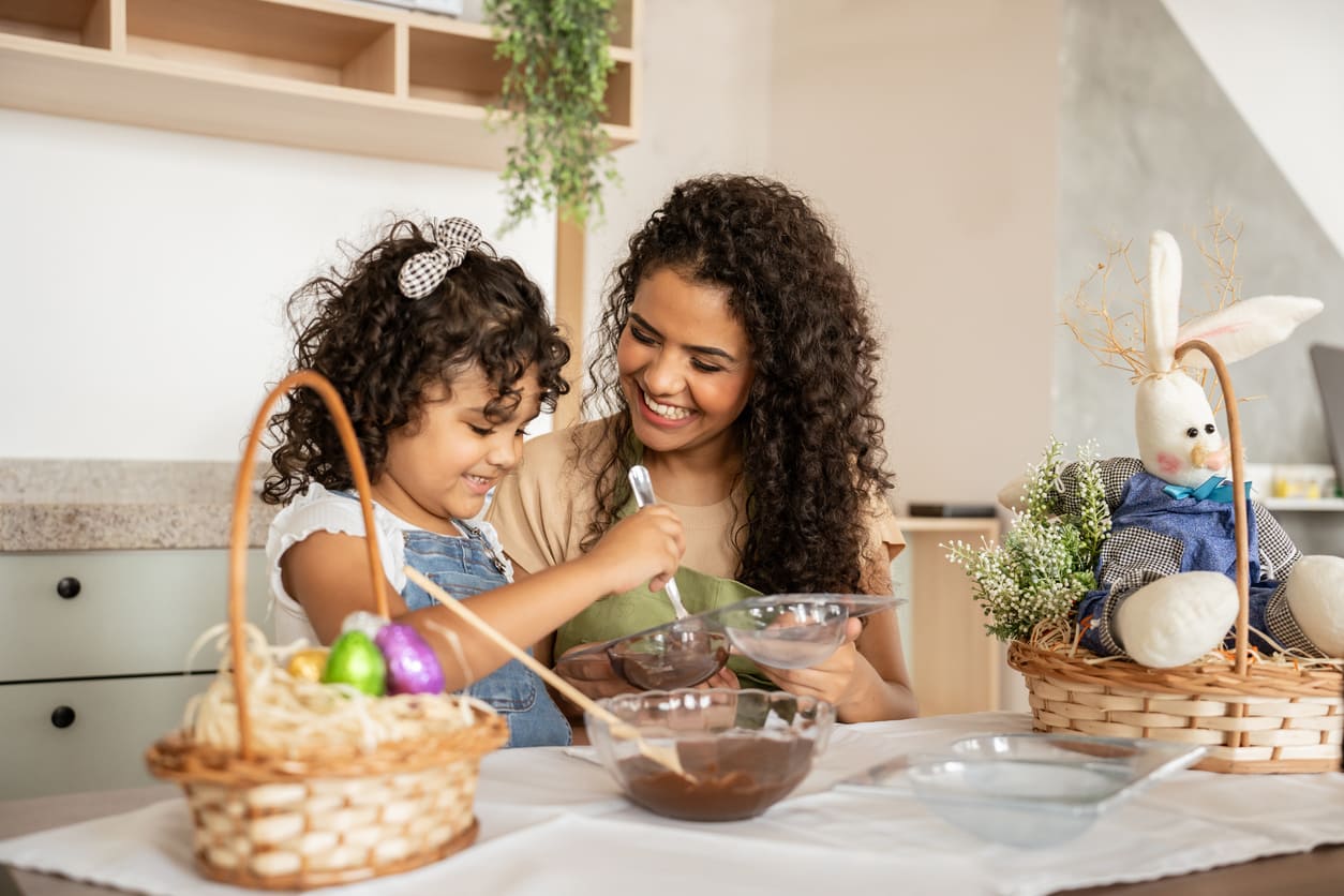 A smiling mother and daughter making chocolate treats together, surrounded by Easter baskets and festive decorations.