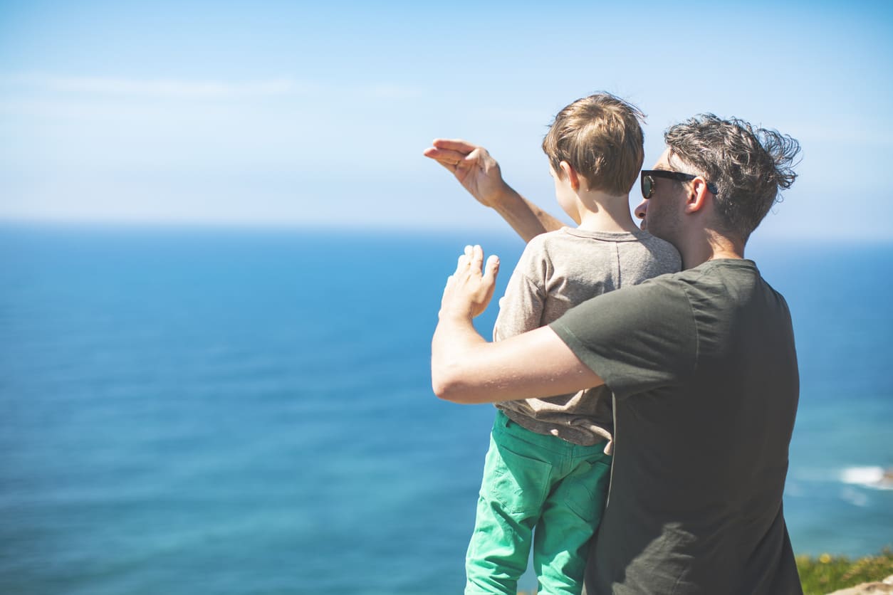 A father holding his child while they look out over the ocean, sharing a peaceful and bonding moment under the clear blue sky.