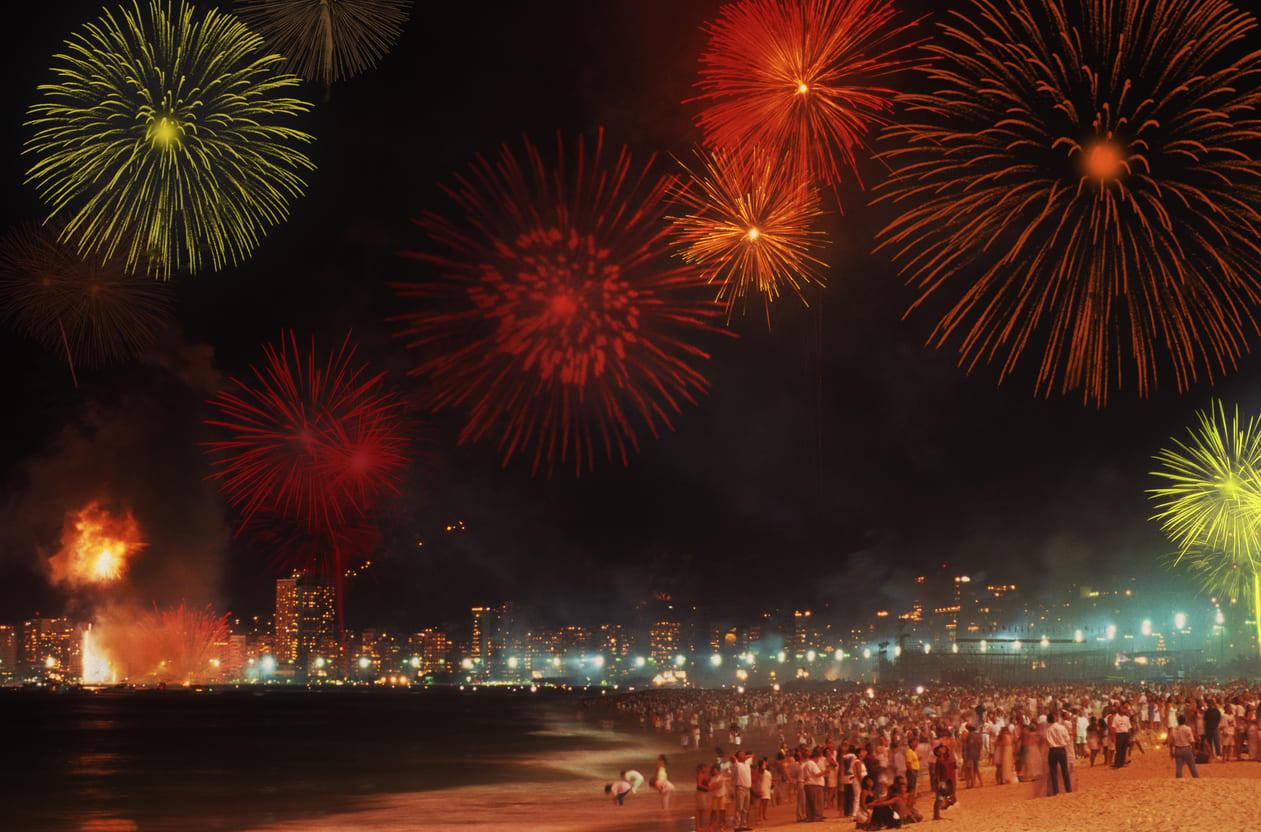 A vibrant night scene on the beach in Rio de Janeiro, with crowds of people dressed in white watching a stunning fireworks display to welcome the New Year.