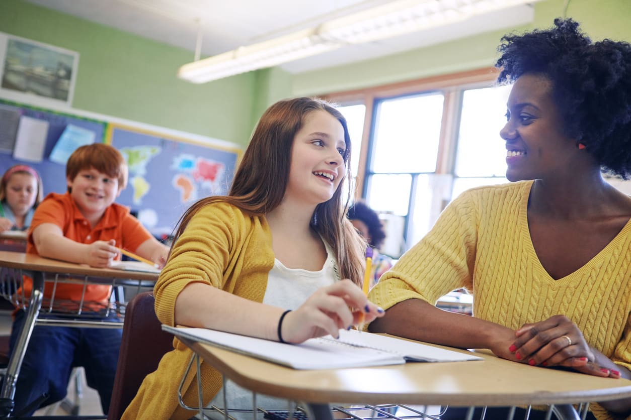 A smiling teacher and student interacting warmly in a classroom, capturing a moment of connection and encouragement.