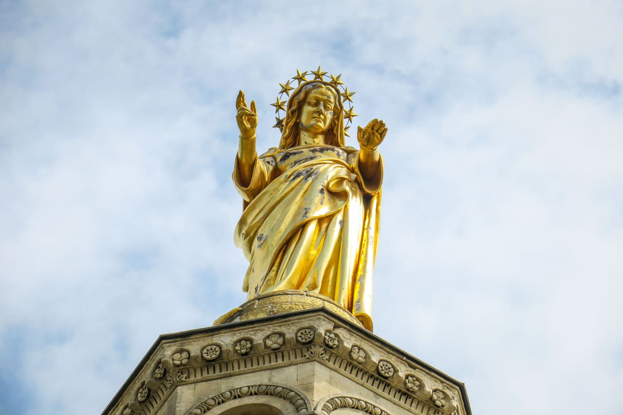 A golden statue of the Virgin Mary, adorned with a halo of stars, stands against the sky, symbolizing her ascension to heaven on Assumption Day.