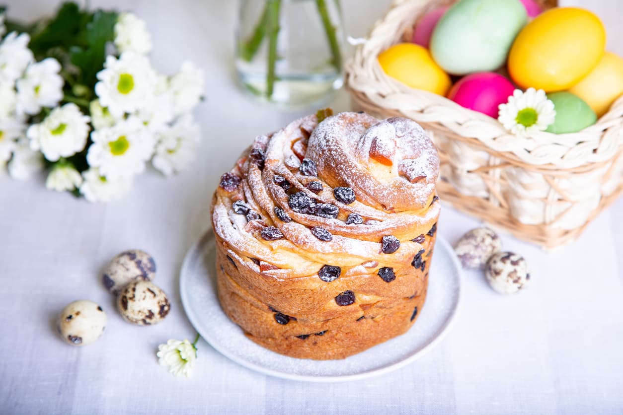 A beautifully arranged Easter scene featuring a festive raisin-studded bread dusted with powdered sugar, a basket of colorful eggs, and delicate spring flowers.