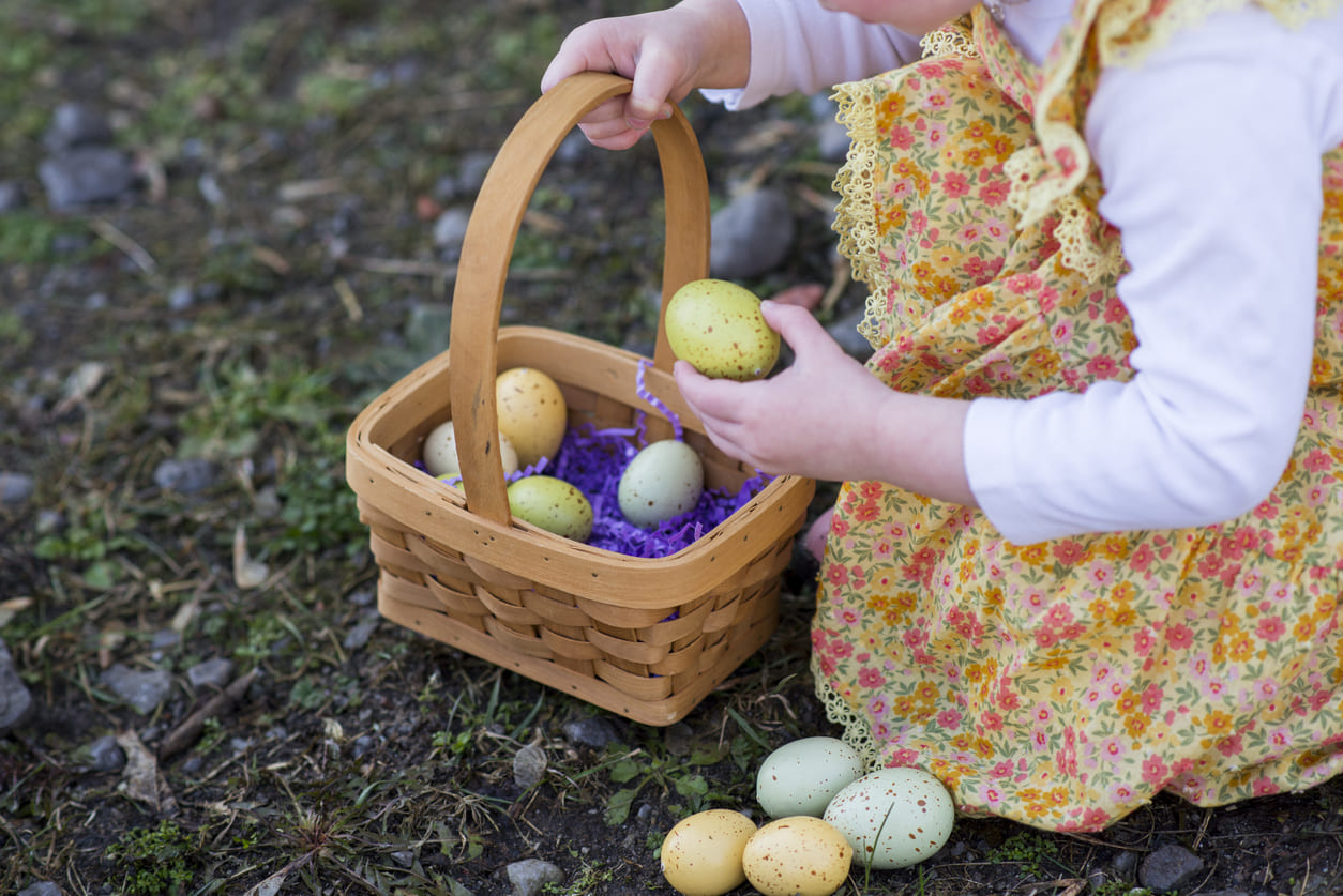 A young child, dressed in a floral outfit, joyfully gathers pastel-colored Easter eggs into a wicker basket during an egg hunt.