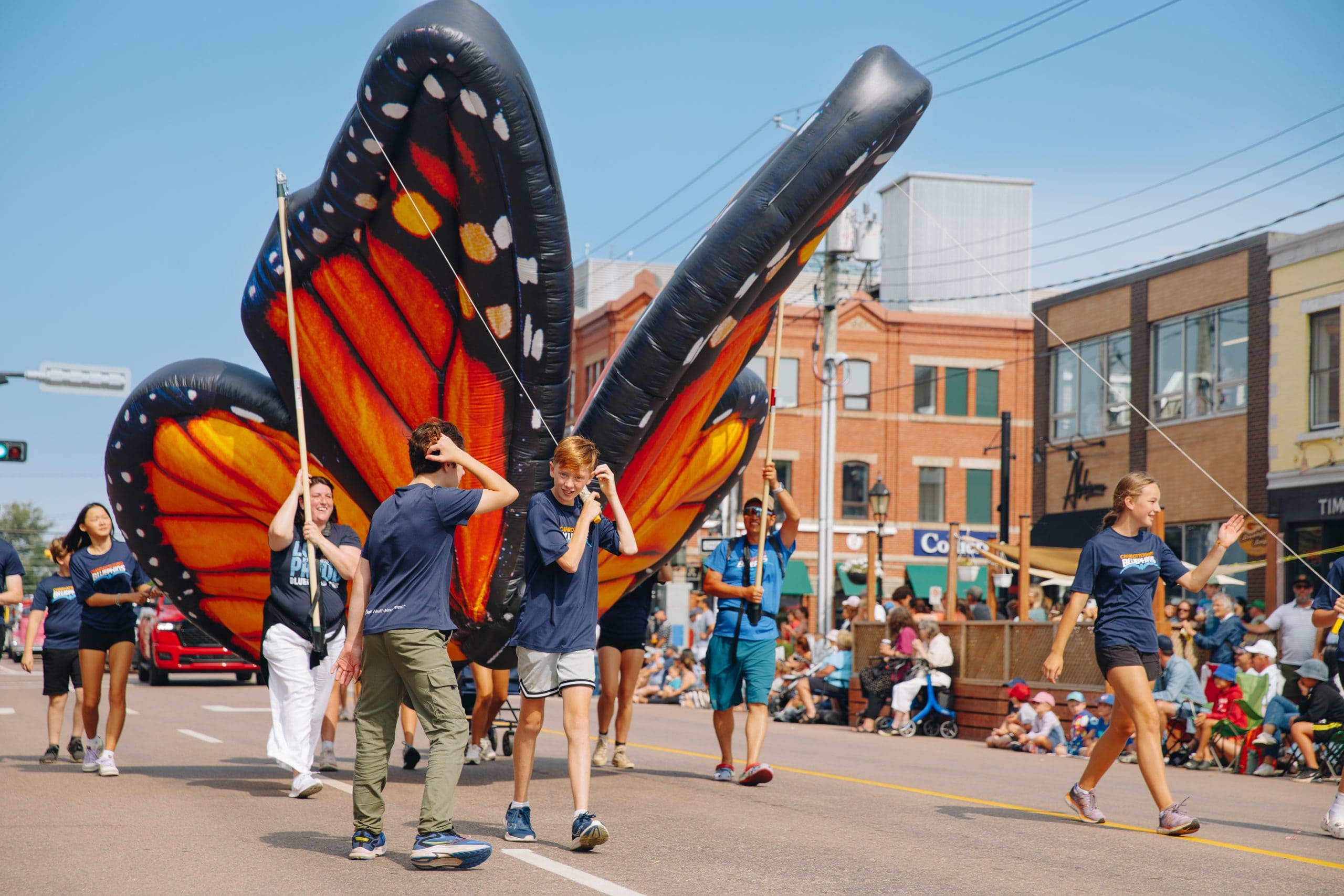Participants in the Gold Cup Parade walk alongside a giant inflatable butterfly float, entertaining crowds in Charlottetown.
