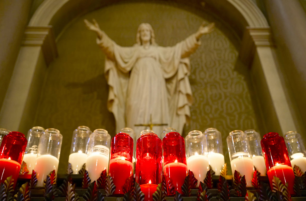 A row of glowing prayer candles in red and white stands before a statue of Jesus, creating a solemn and reflective atmosphere.