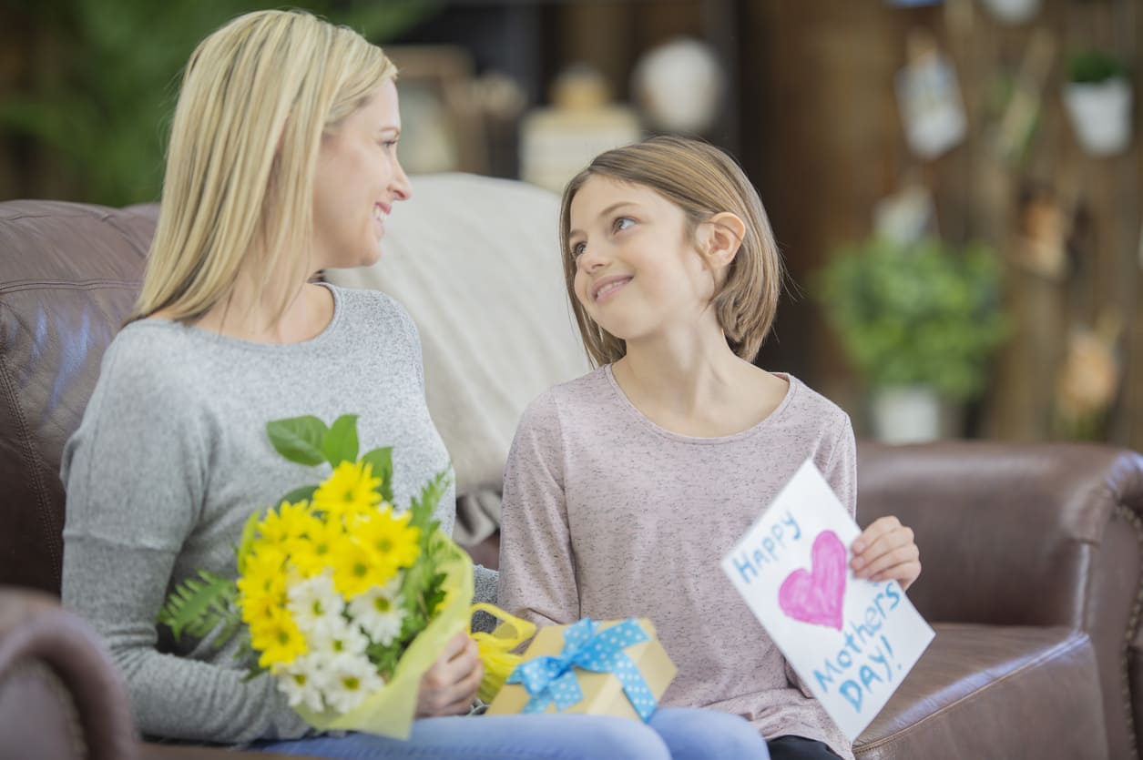 A mother and daughter sharing a warm moment on Mother's Day, with the daughter gifting flowers, a present, and a handmade card.