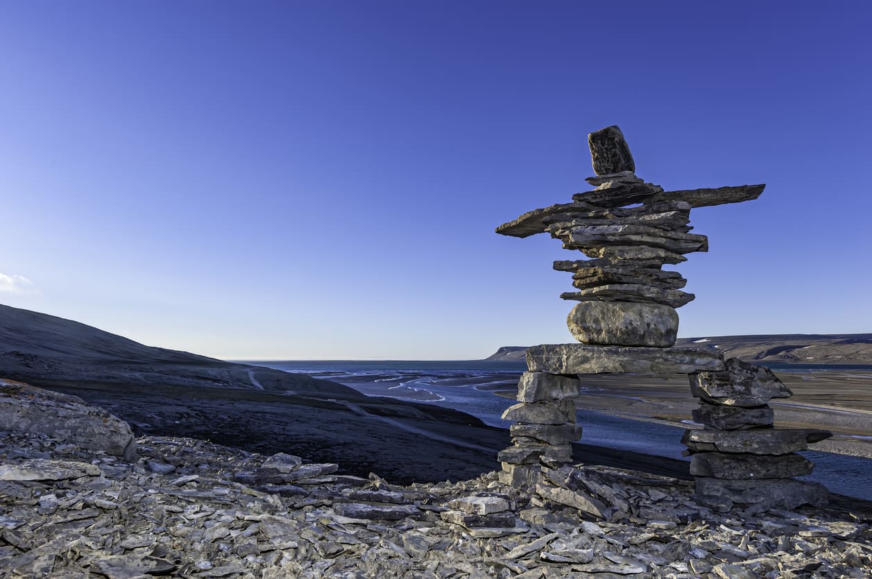 A traditional Inukshuk made of stacked stones stands against the vast Arctic landscape under a clear blue sky.