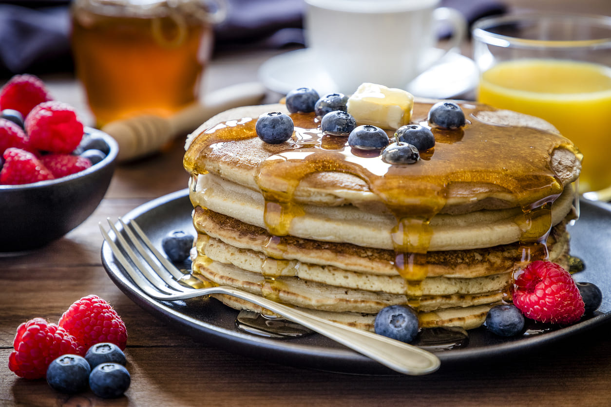 A stack of golden pancakes drizzled with maple syrup, topped with butter and fresh blueberries, with raspberries, honey, and orange juice in the background.