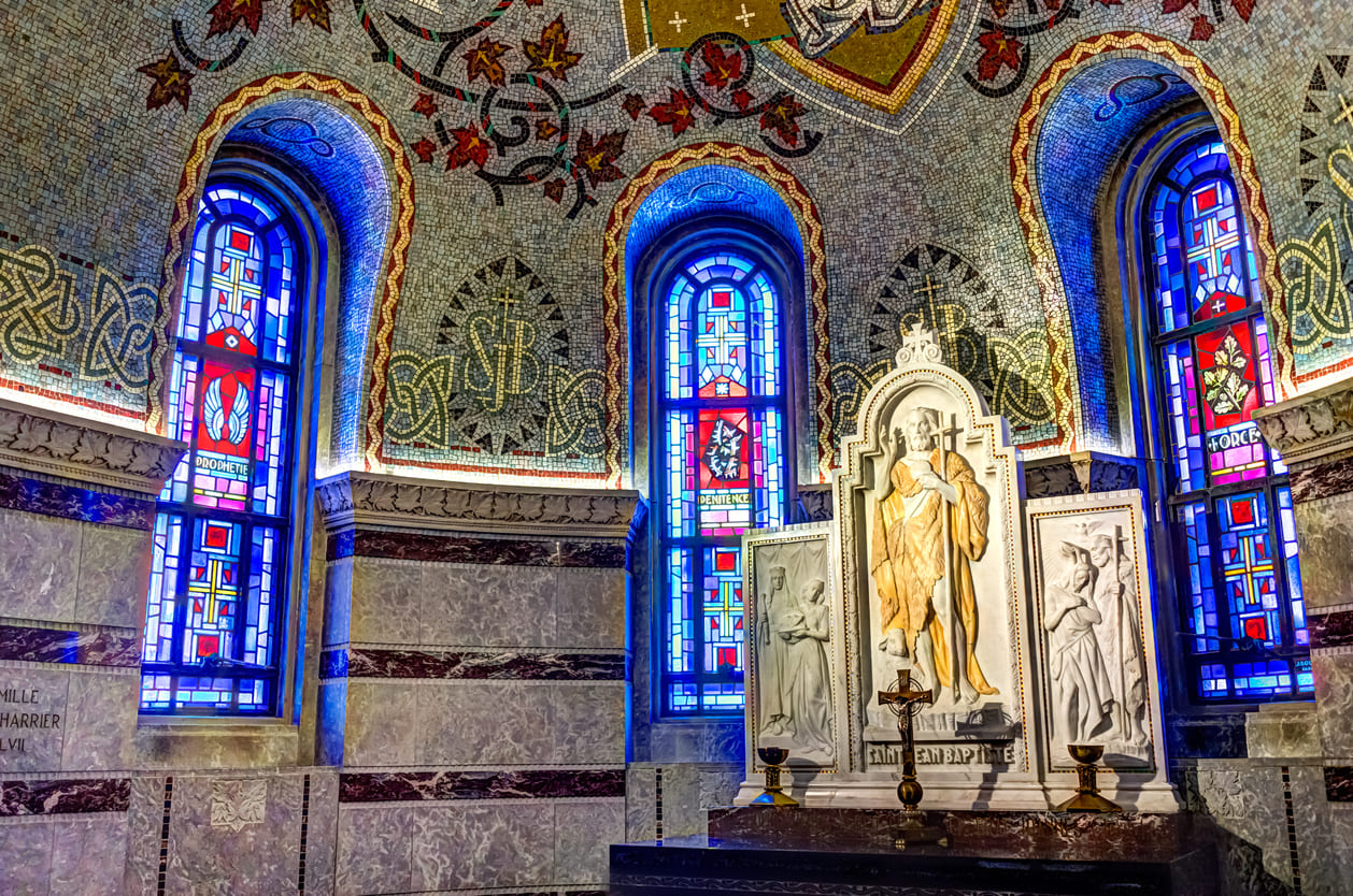 A radiant statue of Saint John the Baptist inside the Basilica of Sainte-Anne-de-Beaupré, Quebec, surrounded by mosaics and stained glass.