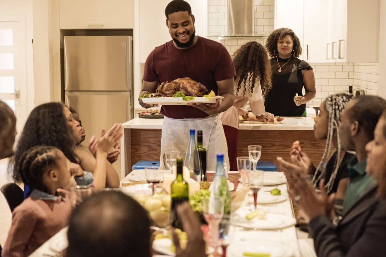 A smiling man presents a roasted turkey to a gathered family, who are clapping and enjoying a warm Thanksgiving celebration in a modern kitchen.