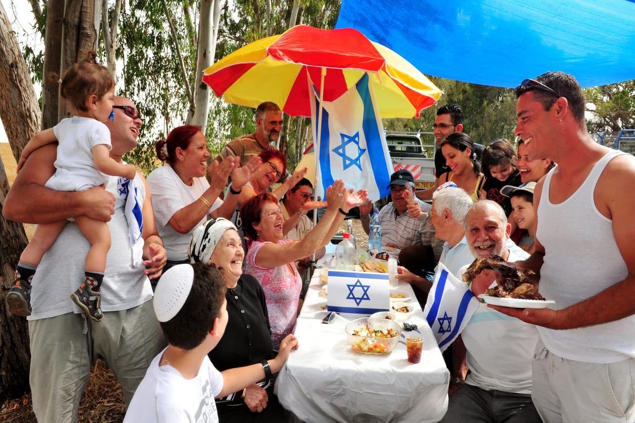 A lively Yom Ha'atzmaut celebration where families gather outdoors, enjoying food, laughter, and Israeli flags in a festive atmosphere.