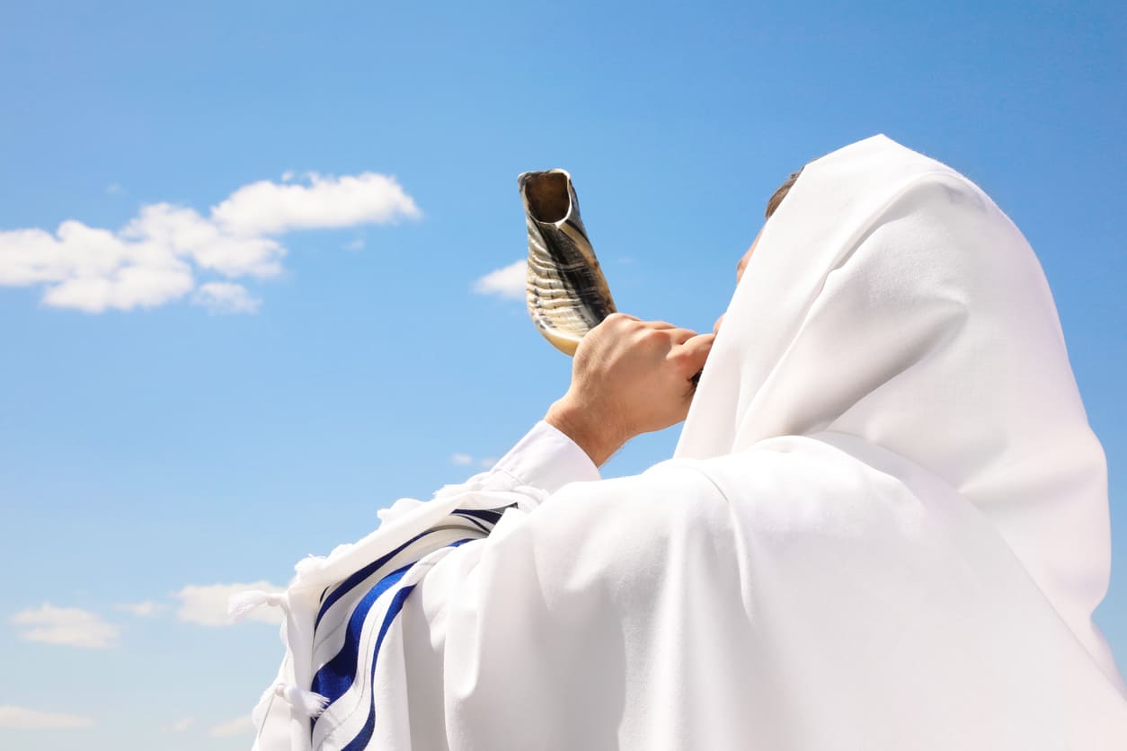 A person wearing a tallit blows the shofar under a clear sky, symbolizing the solemn conclusion of Yom Kippur and spiritual renewal.