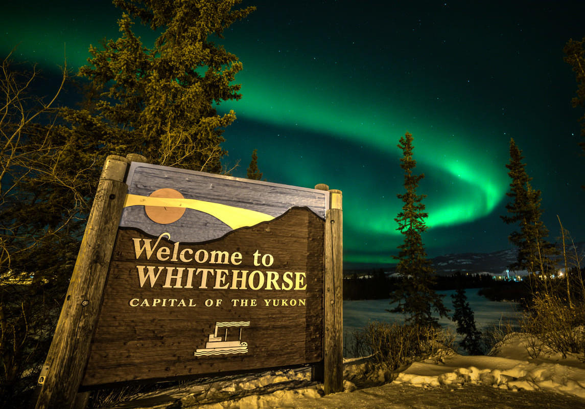 A wooden sign stands against a breathtaking night sky, where the Northern Lights dance above the snowy Yukon landscape.