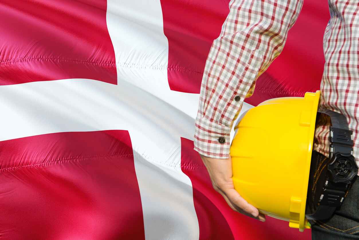 A worker, holding a yellow hard hat, stands to the right against the backdrop of the Danish flag.